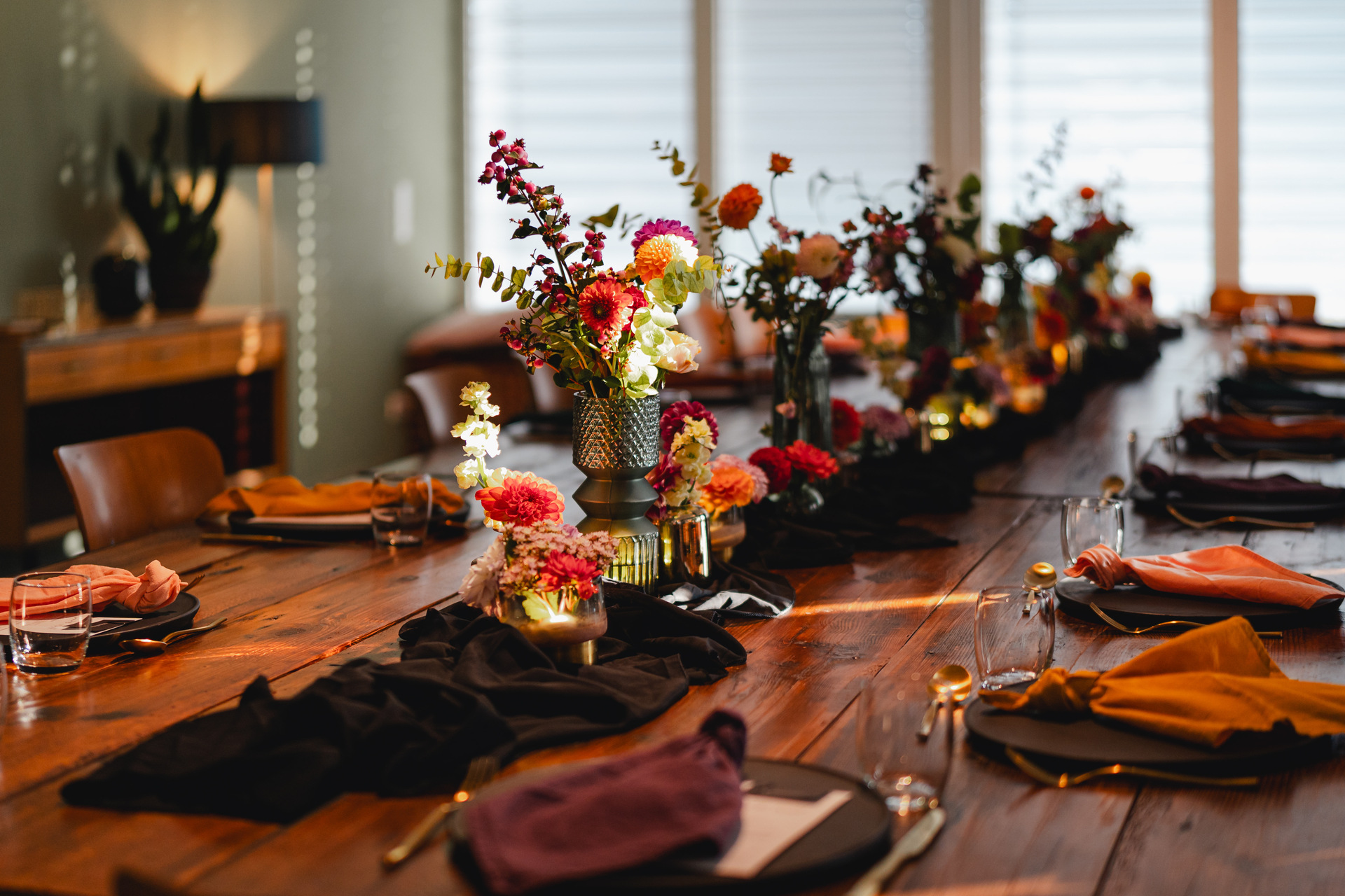 Tischdekoration bestehend aus einem locker liegenden, schwarzen Tischläufer, darauf stehen viele Vasen unterschiedlicher Formen und Farben, mit vielen bunten Blumen, auf einer langen Holztafel