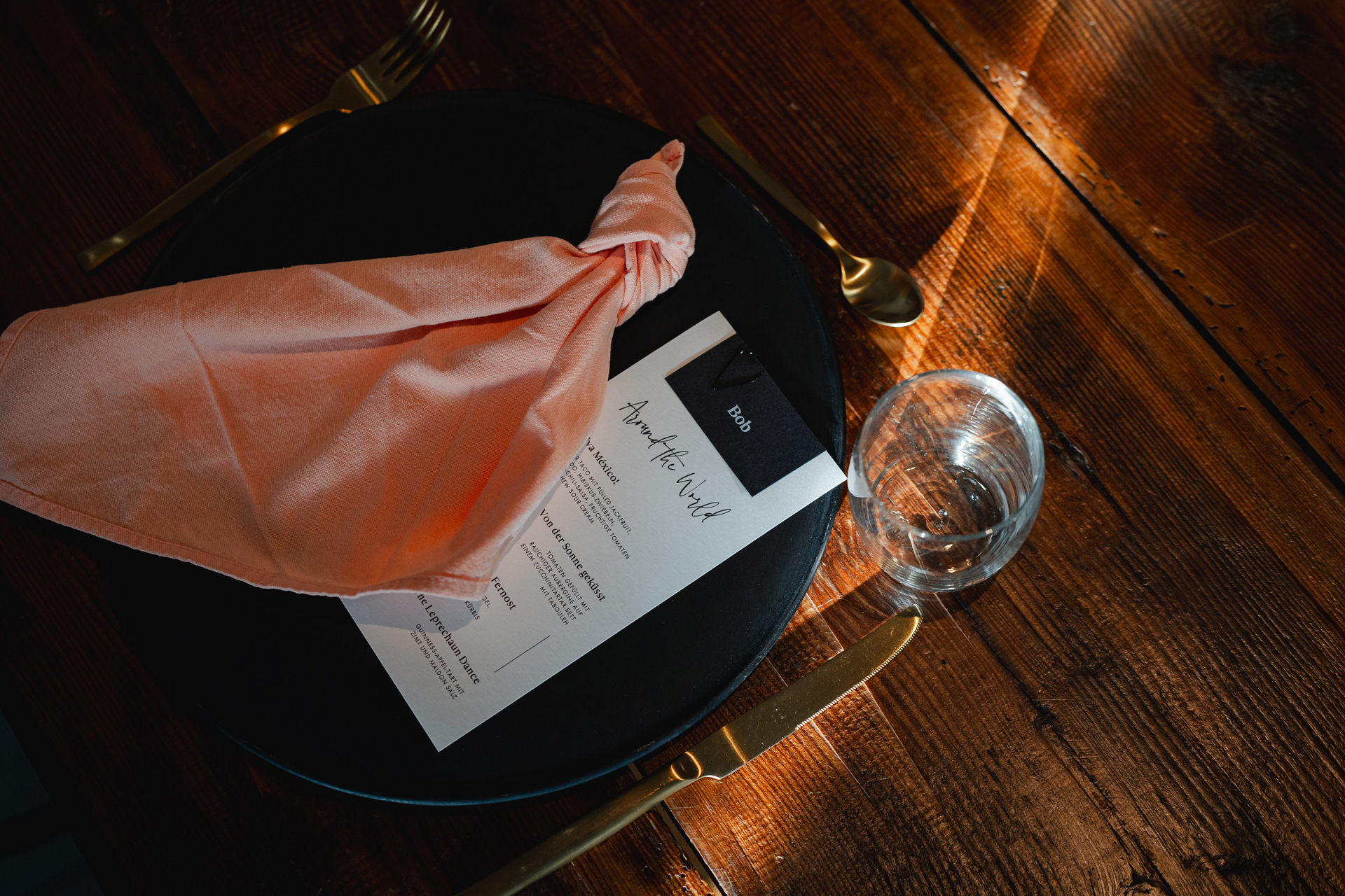 Schlichtes, schwarz-weißes Dinner-Menü, auf einem Teller liegend, daneben eine locker geknotete, lachsfarbene Serviette und goldenes Besteck