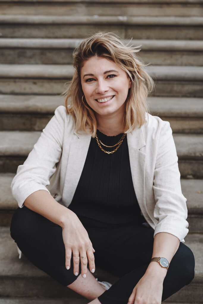 Porträtbild einer jungen lächelnden Frau sitzend auf einer Treppe. Es handelt sich um Sarah Hentgen, Gründerinn von BOLD YES Weddings und Hochzeitsplanerin